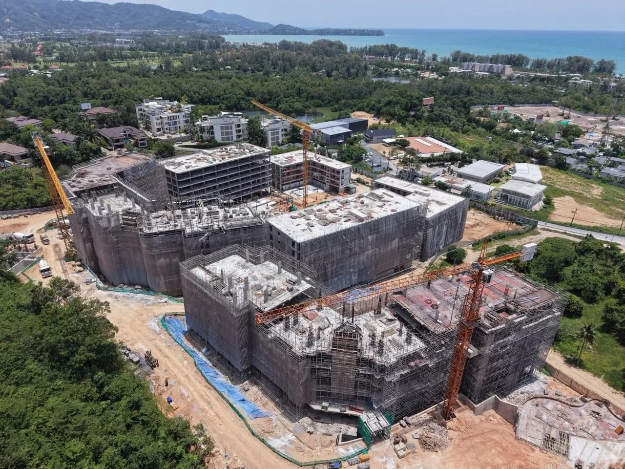 Drone photo, August 2026: blocks E–H topped out under scaffolding, Bang Tao bay behind